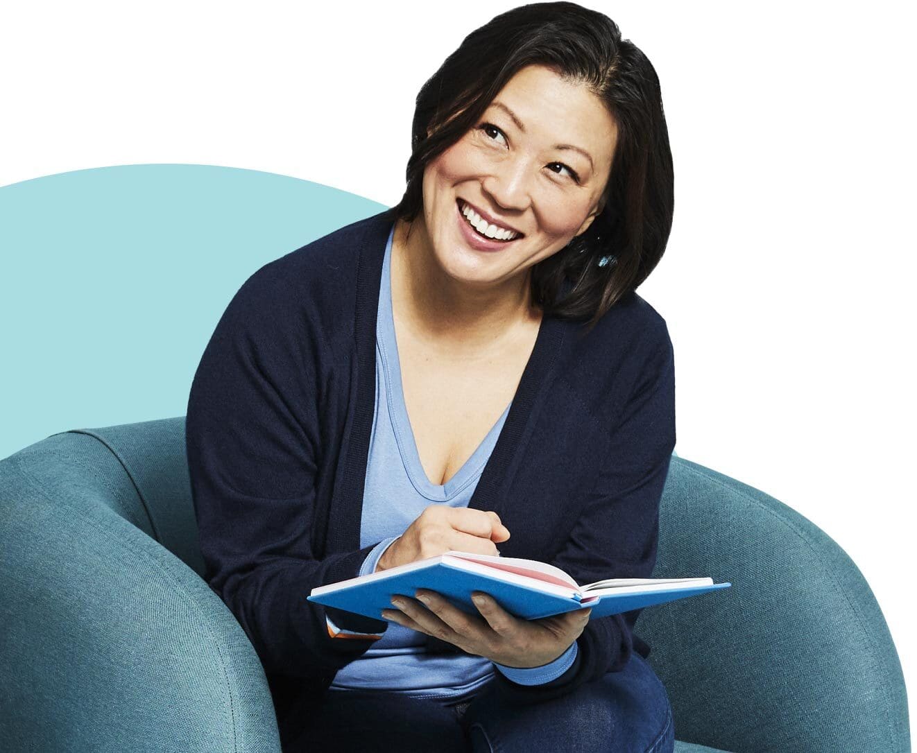 Young Asian woman sits in an armchair, pointing to her notebook and smiling wide